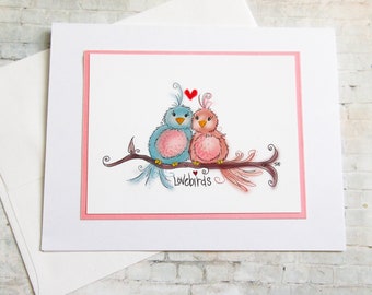May include: A greeting card featuring two cartoon lovebirds perched on a branch, with a red heart above them. The birds are blue and pink, with the word "Lovebirds" written below. The card has a pink border and comes with a white envelope.