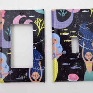 May include: Two black light switch covers with a mermaid theme. The covers feature mermaids with blue hair, playing music, and surrounded by sea life. The background is black with a pattern of fish, seaweed, and bubbles.