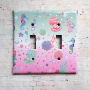Puede incluir: Una placa de interruptor doble con un tema de sirena. La placa presenta un fondo degradado rosa y verde con caballitos de mar, conchas marinas, perlas y estrellas.