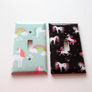 May include: Two light switch covers with a unicorn pattern. One cover is green with white unicorns and rainbows. The other cover is black with white unicorns and stars.
