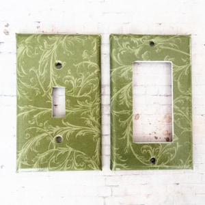 May include: Two rectangular light switch plates featuring a sage green background with a lighter green swirling vine and leaf pattern. One plate has a single vertical toggle switch opening, while the other has a larger rectangular opening.