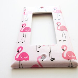 May include: A decorative light switch cover with a repeating pattern of pink flamingos on a pale pink background. The flamingo illustrations have black legs and beaks. The cover is rectangular with a single opening.