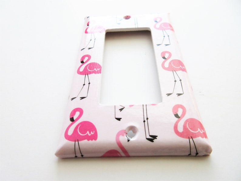 May include: A decorative light switch cover with a repeating pattern of pink flamingos on a pale pink background. The flamingo illustrations have black legs and beaks. The cover is rectangular with a single opening.