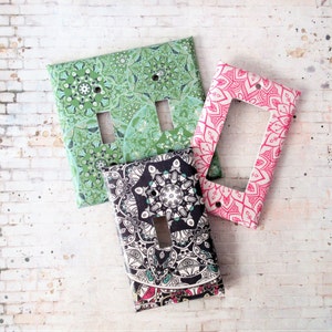 May include: Three decorative light switch covers featuring mandala designs. The first cover is green with a white mandala pattern. The second cover is black with a white mandala pattern. The third cover is pink with a white mandala pattern.