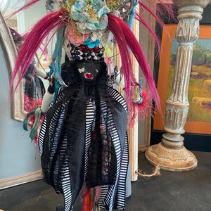 May include: A whimsical doll with pink hair, a black and white striped dress, and a colorful floral headpiece. The doll is standing in front of a mirror and a decorative pillar.