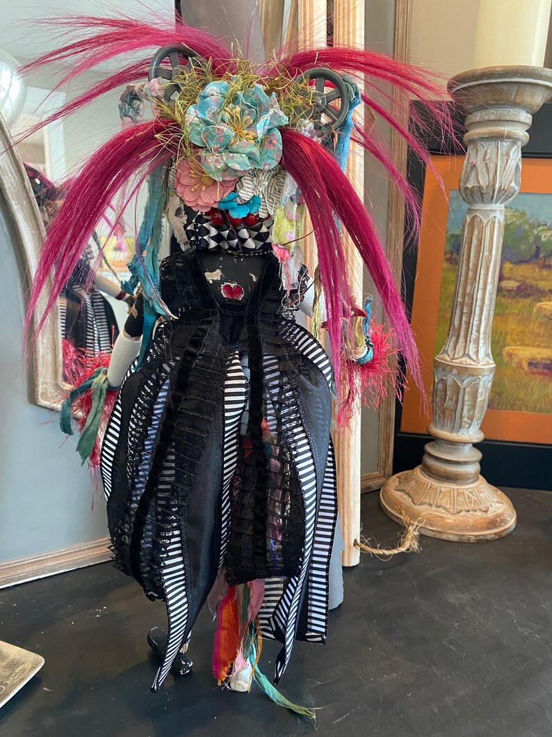 May include: A whimsical doll with pink hair, a black and white striped dress, and a colorful floral headpiece. The doll is standing in front of a mirror and a decorative pillar.