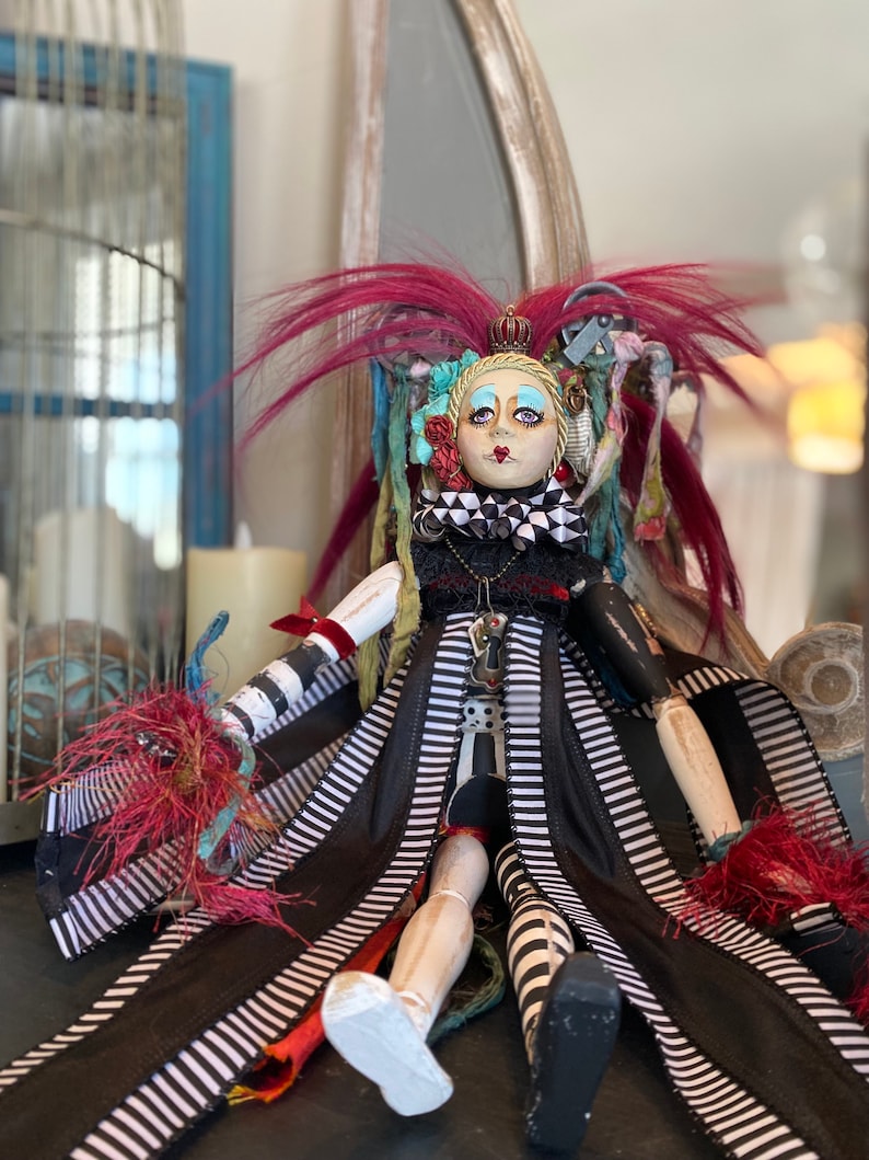 May include: A whimsical doll with a black and white striped dress, red hair, and a crown. The doll has a painted face with blue eyes and a black and white checkered collar.