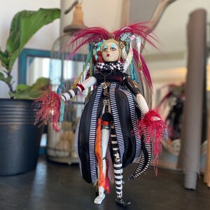 May include: A whimsical doll with a black and white striped dress, red and white feathered accents, and a painted face. The doll has a crown and is wearing a necklace.