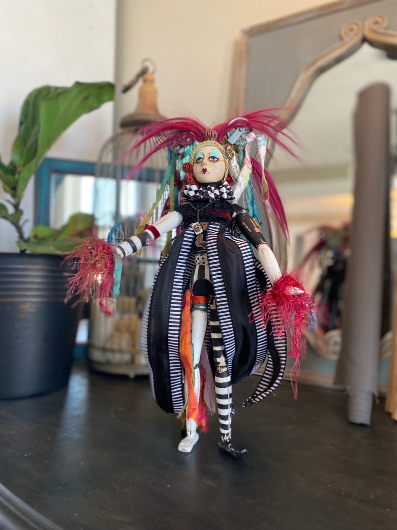 May include: A whimsical doll with a black and white striped dress, red and white feathered accents, and a painted face. The doll has a crown and is wearing a necklace.