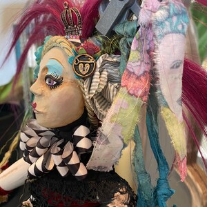 May include: A handmade doll with a black and white checkered bow, a crown, and a heart-shaped key charm. The doll has pink and blue hair and is wearing a black and white striped dress.