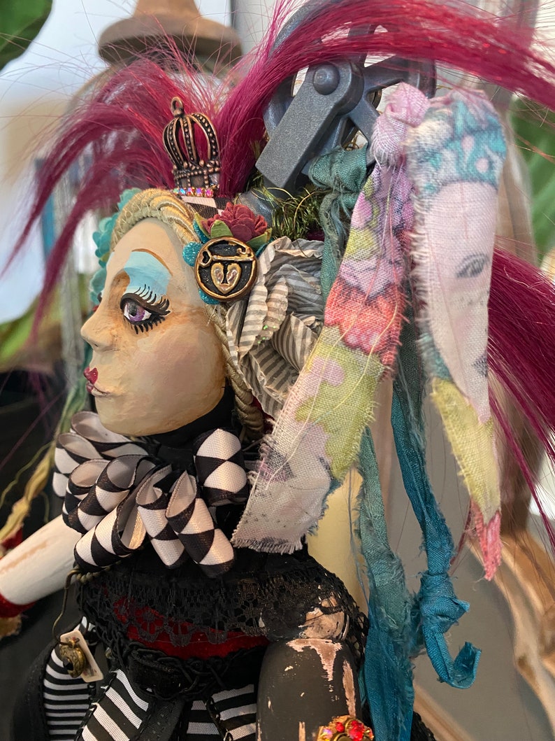 May include: A handmade doll with a black and white checkered bow, a crown, and a heart-shaped key charm. The doll has pink and blue hair and is wearing a black and white striped dress.