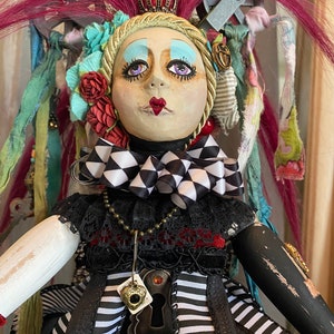 May include: A whimsical doll with a black and white checkered bow, a crown, and a keyhole pendant. The doll has pink hair, blue eyeshadow, and red lips. The doll is wearing a black and white striped dress with a black lace overlay.