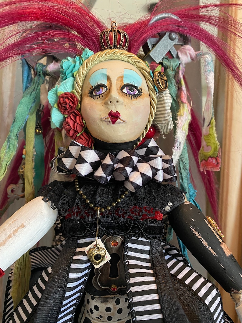 May include: A whimsical doll with a black and white checkered bow, a crown, and a keyhole pendant. The doll has pink hair, blue eyeshadow, and red lips. The doll is wearing a black and white striped dress with a black lace overlay.