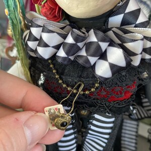 May include: A doll wearing a black and white checkered bow, a red and black lace top, and a black and white striped skirt. The doll is holding a small card with a heart-shaped charm attached to a chain.