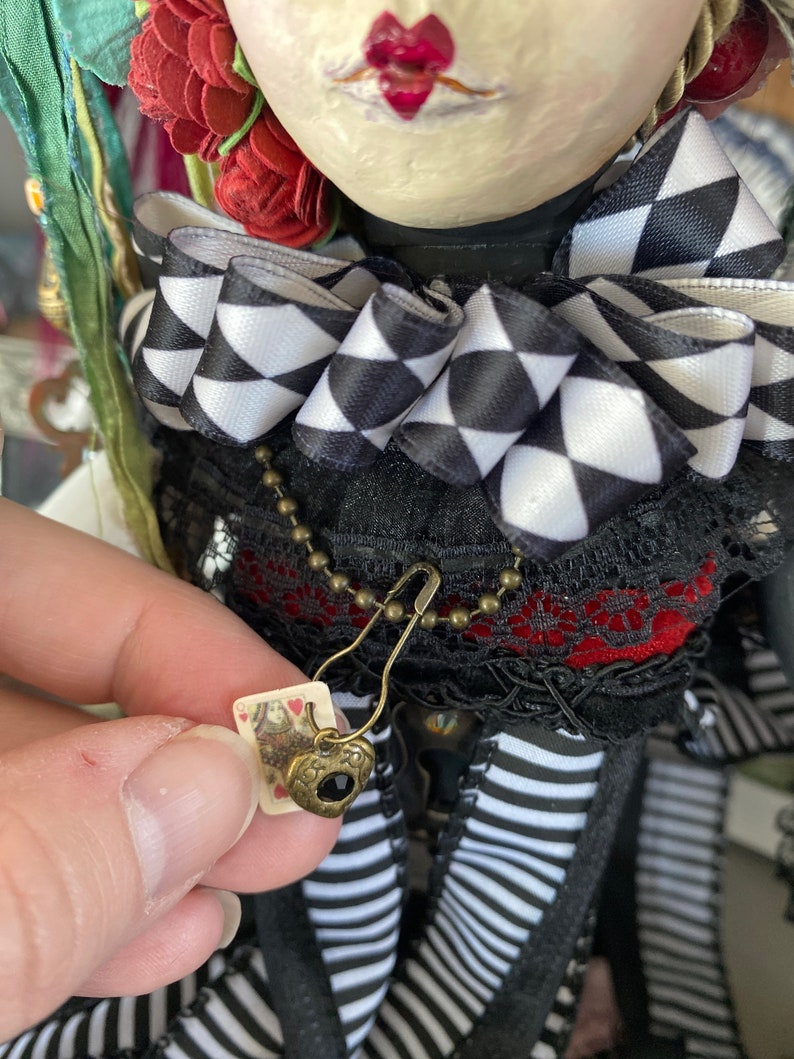 May include: A doll wearing a black and white checkered bow, a red and black lace top, and a black and white striped skirt. The doll is holding a small card with a heart-shaped charm attached to a chain.