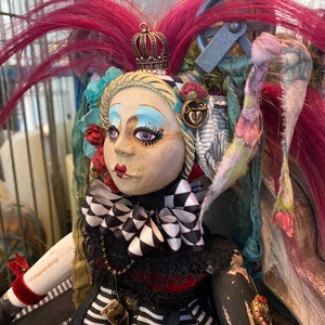 May include: A handmade doll with a black and white striped dress, a black and white checkered bow, and a crown. The doll has pink hair, blue eyeshadow, and red lips. The doll is wearing a necklace with a charm.