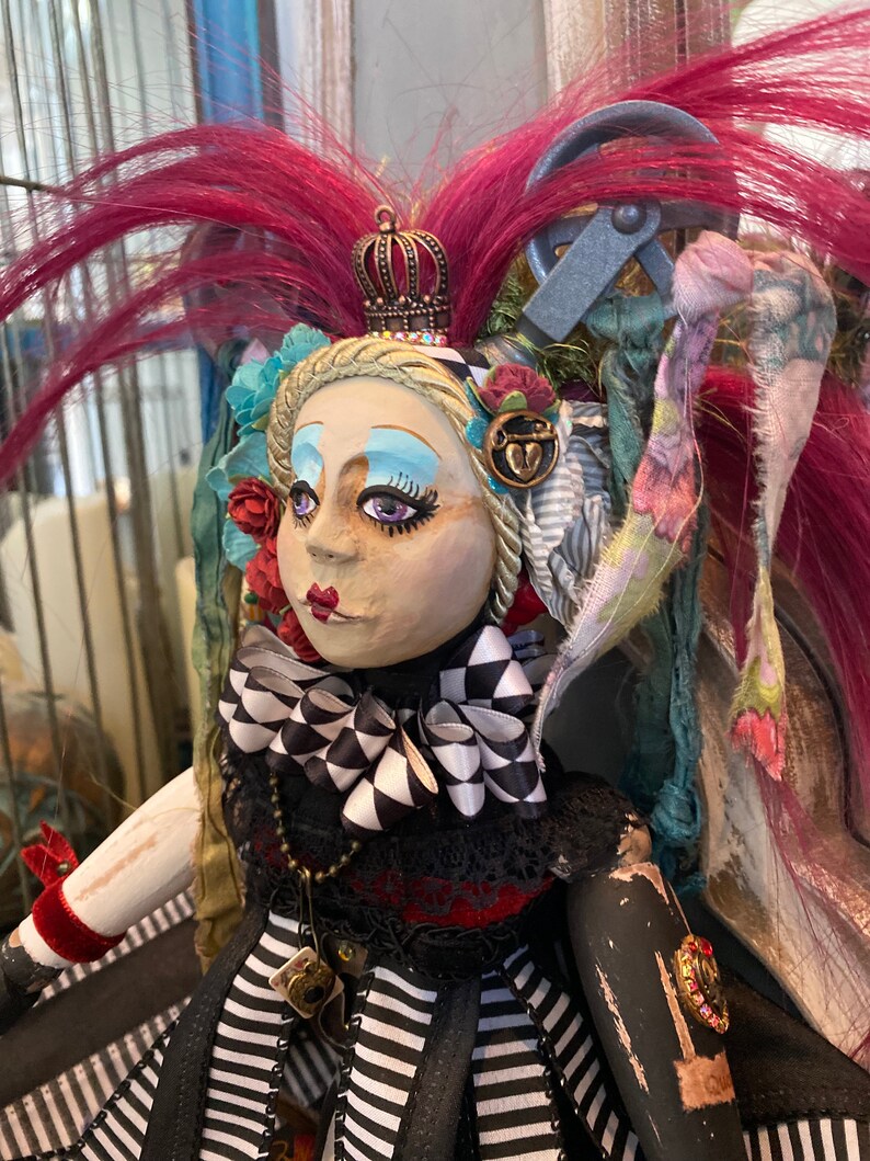 May include: A handmade doll with a black and white striped dress, a black and white checkered bow, and a crown. The doll has pink hair, blue eyeshadow, and red lips. The doll is wearing a necklace with a charm.