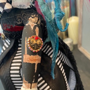 May include: A black and white striped fabric doll with a black lace trim and a red heart-shaped rhinestone button with a gold filigree design. The doll's arm is painted black with white accents and the word "Queenie" written on it.