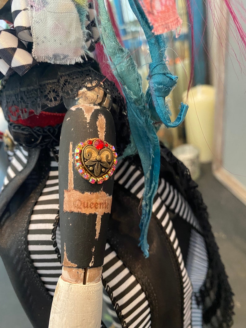 May include: A black and white striped fabric doll with a black lace trim and a red heart-shaped rhinestone button with a gold filigree design. The doll's arm is painted black with white accents and the word "Queenie" written on it.