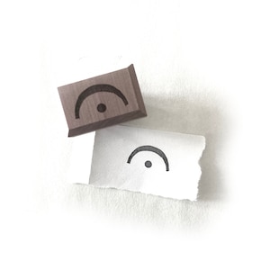 May include: A wooden stamp with a curved line and a dot, and a matching printed image on torn paper. The stamp is brown, and the printed image is black on white. The design is a musical rest symbol.