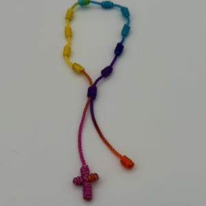 May include: A handmade rosary with a rainbow color scheme. The rosary features knotted beads in a spectrum of colors, including yellow, green, blue, purple, and pink. A small, woven cross hangs at the bottom. The rosary is made of a soft, flexible material.