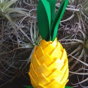 Könnte beinhalten: Eine gelbe Stoff-Ananas mit grünen Stoffblättern. Die Ananas besteht aus gefaltetem Stoff und hat eine strukturierte Oberfläche.