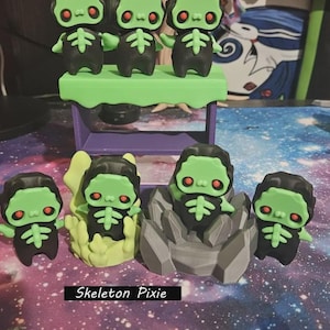 May include: A collection of 3D-printed Skeleton Pixie figurines. The figures have green faces, red eyes, and black bodies with a skeleton design. Some are on a green platform, others in a rock-like structure, and one in a yellow nest.