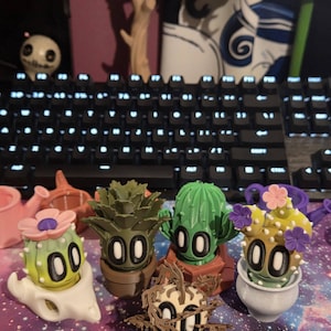 May include: A collection of 3D-printed cactus figurines with cartoon faces and various colors. The cacti are in pots or on stands, with some featuring flower accents. A keyboard and other decorative items are in the background.