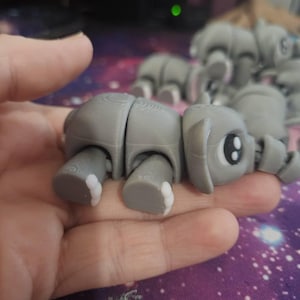 May include: A gray articulated elephant toy with white accents on its feet is held in a hand. The elephant has large eyes and a trunk. Other similar toys are visible in the background.