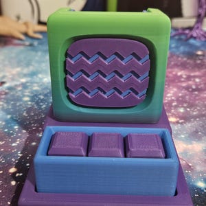 May include: A 3D-printed, multi-colored object featuring a green top, a purple and blue zig-zag design, and a blue tray holding three purple square objects. The base is purple. The design is geometric and modern.