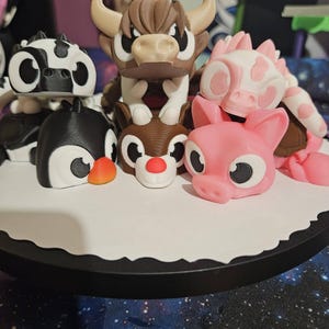 May include: A collection of colorful animal figurines, including a cow, penguin, reindeer, pig, and a horned bull. The figurines are arranged on a white, scalloped-edge base with a black border. The animals are in various colors, including black, white, brown, pink, and orange.