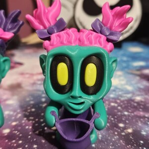 May include: A teal figurine with pink hair, purple bows, and yellow eyes. The figurine is holding a purple bucket. The background includes a Jack Skellington figure and a galaxy-themed surface.
