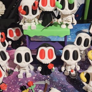 May include: A collection of white skeleton figurines with black and red accents. Some skeletons are holding a scythe, a rose, or a knife. The figurines are arranged on a surface with a galaxy-themed background. The figurines have different colored accents.