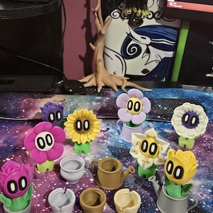 May include: A collection of whimsical flower figurines with cartoon-like faces and various colors, including pink, yellow, and white. Each flower has a green stem and is paired with a small pot or watering can. The figurines are arranged on a galaxy-themed surface.