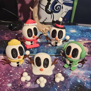 May include: A collection of whimsical snowman figurines with various accessories, including hats, scarves, and earmuffs. The snowmen are white with black oval eyes and mouths, and brown twig arms. Some hold snowballs. The figurines are on a galaxy-themed surface.
