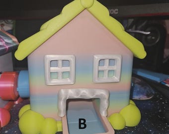 May include: A 3D-printed house-shaped container in pastel colors. The roof and decorative accents are lime green. The house has two square windows and a rectangular opening with the letter "B".