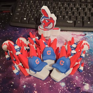 May include: A collection of 3D-printed baseball-themed figurines. The figurines include baseballs, baseball caps with a "C" logo, and a white foam finger with a Native American caricature. The color scheme is red, white, and blue.