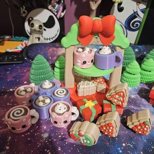 May include: A whimsical Christmas-themed display featuring miniature mugs of hot chocolate, gingerbread houses, and wrapped gifts. The mugs are pink and purple, some with whipped cream and marshmallows. The scene includes a red bow and green trees.