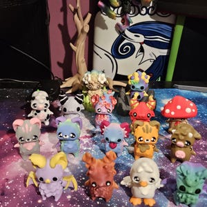 May include: A collection of colorful, small figurines with various animal-like and fantasy creature designs. The figurines feature a range of colors, including pink, blue, orange, and green, with unique details and expressions. Some have wings, horns, or mushroom caps.