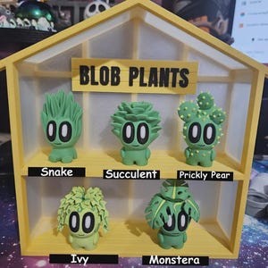 May include: A yellow display case with the text "BLOB PLANTS" houses five green, cartoon-like plant figures. Each figure has large, round eyes and a unique plant-inspired design, labeled "Snake," "Succulent," "Prickly Pear," "Ivy," and "Monstera."