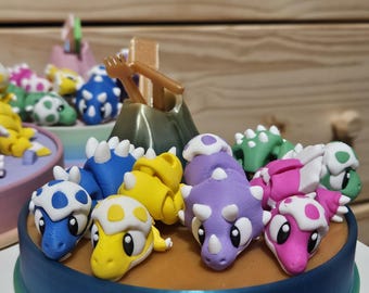May include: A colorful, handcrafted cake featuring multiple dinosaur figurines in various colors, including blue, yellow, purple, and pink. The cake has a blue base and a brown surface, with a volcano-shaped decoration.