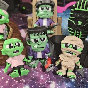May include: A collection of 3D-printed monster figurines, including Frankenstein's monster, a mummy, and zombies. The Frankenstein figure is holding a tool. The figures are green, purple, and tan, with black and white accents. They are on a purple and blue surface.