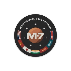 May include: A black and white circular patch with the text "International Mars Program" around the edge. In the center is a red planet with the text "M-7" in white. There are flags from different countries around the planet, including the United States, the Soviet Union, North Korea, Japan, India, and the European Space Agency.