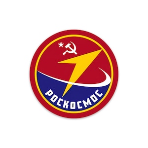 Puede incluir: Un parche circular rojo, blanco y azul con un rayo amarillo y el texto "РОСКОСМОС" en escritura cirílica. El parche también presenta una estrella roja y una hoz y un martillo.