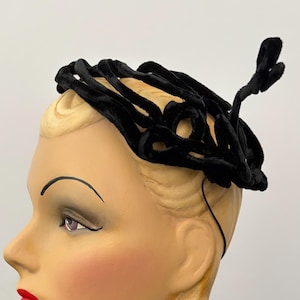 Op de afbeelding: Een zwarte fluwelen fascinator met een reeks onderling verbonden, geluste fluwelen linten in een cirkelvormig patroon, bevestigd aan een dunne, donkere haarband, met een gestructureerd en driedimensionaal ontwerp.