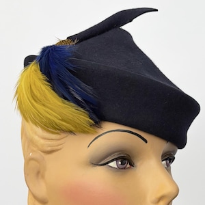 Op de afbeelding: Een marineblauwe hoed met een unieke vorm, versierd met gele en blauwe veren. De hoed wordt getoond op een mannequinhoofd met blond haar, rode lippenstift en blozende wangen. De hoed heeft een kleine, puntige rand.