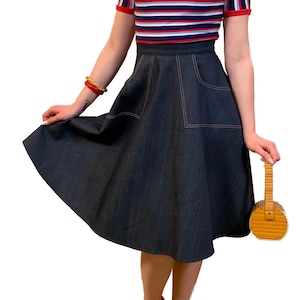 Op de afbeelding: Een vrouw in een nautische outfit. De outfit bestaat uit een gestreepte top met rode bies, een spijkerbroekrok en rode schoenen. Ze houdt een kleine, ronde houten tas vast. Een bloem zit in haar haar.