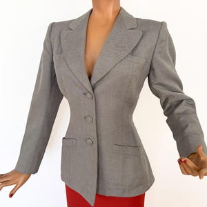 Puede incluir: Una blazer gris con un corte entallado, con cierre de dos botones y dos bolsillos delanteros. La blazer se exhibe en un maniqu&iacute; con una falda roja. El maniqu&iacute; tiene un peinado vintage y mira hacia arriba.