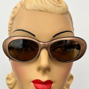 Op de afbeelding: Een paar ovale zonnebrillen met een bruin montuur en bruine glazen. De zonnebril zit op het gezicht van een mannequin.