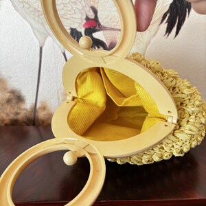 May include: A vintage heart-shaped handbag with a woven straw exterior and a yellow fabric interior. The bag features a cream-colored frame and handle, with a clasp closure. The bag is open, revealing the bright yellow lining.